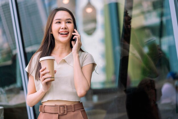 La programadora nómada digital relajada casual asiática mira una conversación en un teléfono inteligente mientras camina en la ciudad con una taza de café sosteniendo una sonrisa alegre y positiva que cambia el estilo de vida