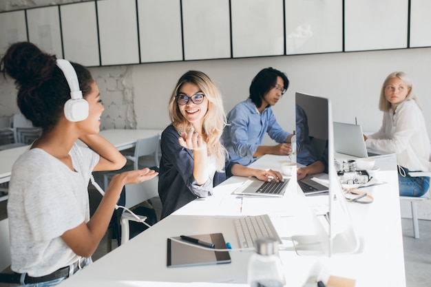 Programadoras web con estilo hablando sobre el trabajo mientras pasan tiempo en la oficina. Retrato de interior de mujer africana en auriculares y trabajador asiático usando computadoras.