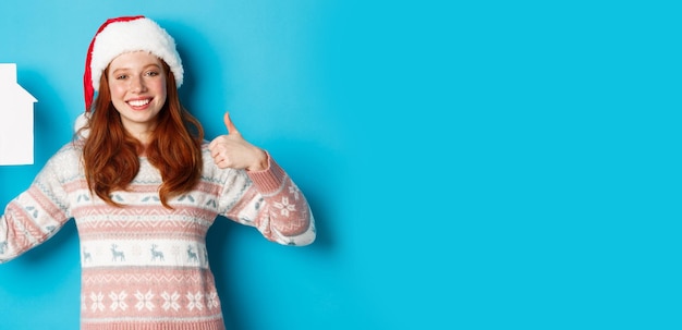 Foto gratuita promociones de vacaciones y concepto de bienes raíces modelo femenino satisfecho con cabello ondulado rojo con sombrero de santa a