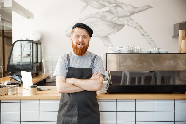 Propietario de negocio de café Concepto Retrato de feliz joven barista caucásico barbudo en delantal con mirada confiada y sonriendo a la cámara en el mostrador de la cafetería