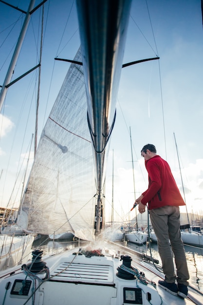 El propietario de un velero o un navegante utiliza una manguera para lavar el agua salada de la cubierta del yate cuando está atracado o estacionado en la marina al atardecer