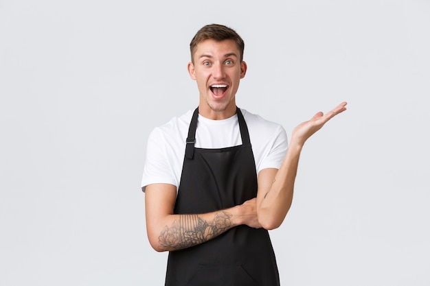 Propietarios de pequeñas empresas, cafetería y concepto de personal. Aliviado y feliz sonriente barista guapo regocijándose, levantando la mano y mirando emocionado, contando grandes noticias, publicidad.