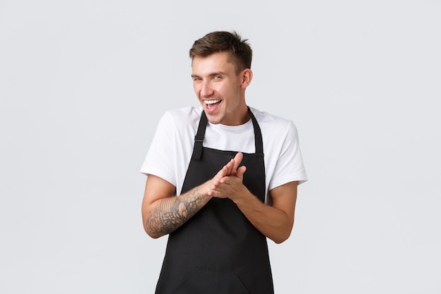 Propietarios de pequeñas empresas, cafetería y concepto de personal. Barista divertido, astuto e inteligente con delantal negro, trabajador del café frotándose las manos como si sintiera placer o satisfacción por el plan cumplido, sonriendo maliciosamente.