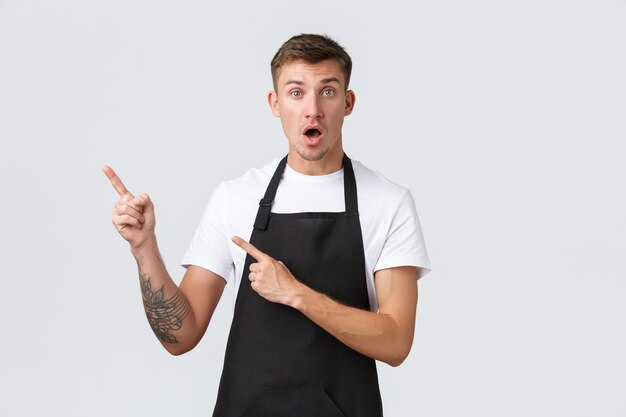 Propietarios de pequeñas empresas, cafetería y concepto de personal. Chico guapo asombrado e impresionado, camarero en la cafetería o restaurante señalando con el dedo la esquina superior izquierda, mirando con la boca abierta a la cámara