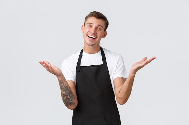 Foto gratuita propietarios de pequeñas empresas, cafetería y concepto de personal. chico guapo de aspecto amistoso que parece encantado y feliz, invitando a los invitados a la cafetería, barista que trabaja a tiempo parcial en el restaurante