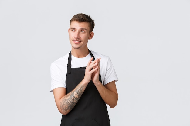 Propietarios de pequeñas empresas, cafetería y concepto de personal. Creativo e inteligente barista astuto guapo, camarero o empleado de café en delantal negro, dedos de campanario y sonrisa, tiene una idea genial, fondo blanco.