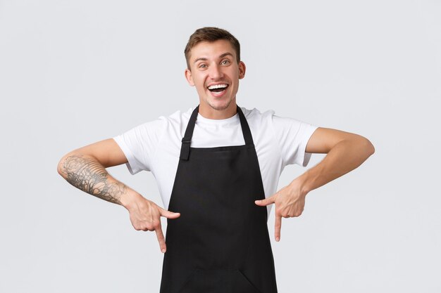 Propietarios de pequeñas empresas, cafetería y concepto de personal. Feliz y amigable barista, camarero con delantal negro apuntando con el dedo hacia abajo, chico que trabaja en la cafetería y muestra una promoción o un anuncio especial