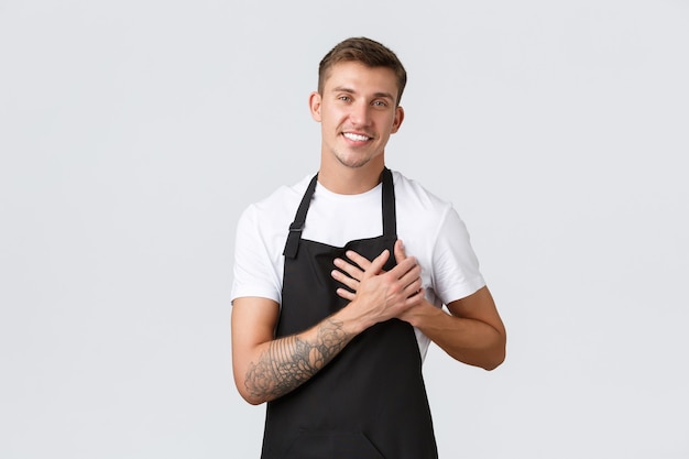 Propietarios de pequeñas empresas, cafetería y concepto de personal. Feliz chico rubio guapo con delantal negro, barista o camarero sosteniendo la mano en el corazón y sonriendo, aprecia a los invitados del café, ama a cada cliente