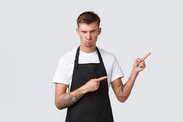 Propietarios de pequeñas empresas, cafetería y concepto de personal. Moody y escéptico joven empleado de café, barista odia el mostrador de limpieza, apuntando hacia la esquina superior derecha y mirando a la cámara enojada, haciendo muecas