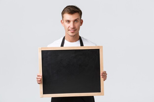 Propietarios de pequeñas empresas minoristas, concepto de empleados de cafetería y restaurante. Vendedor atractivo y amistoso anunciar algo a los clientes, sosteniendo el tablero sin carteles, sonriendo feliz, fondo blanco