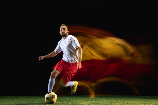 Foto gratuita proponer. jugador de fútbol o fútbol masculino caucásico joven en ropa deportiva y botas pateando la pelota para el gol en luz mixta en la pared oscura. concepto de estilo de vida saludable, deporte profesional, afición.