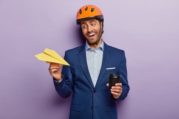 Próspero hombre de negocios feliz posando en elegante traje y casco rojo en la oficina