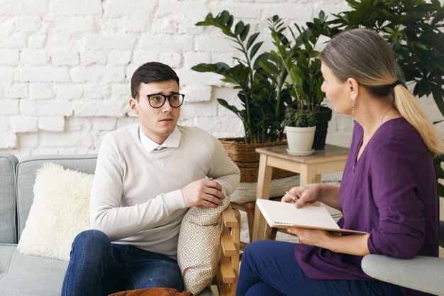 Psicoterapeuta o consejero de la mujer mayor que escribe algo en el cuaderno durante la sesión de terapia con el varón joven deprimido frustrado en espectáculos. Psicología, asesoramiento y salud mental