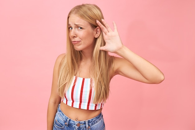 ¿puede hablar más alto? retrato de moda linda joven mujer en top de verano y jeans frunciendo el ceño, sosteniendo la mano en su oreja, teniendo problemas de audición debido al ruido