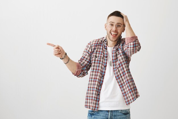 ¿Puedes creer que vi a una celebridad por ahí Foto de estudio de un compañero de trabajo barbudo, asombrado y emocionado, apuntando a la izquierda con el dedo índice sonriendo y rascándose la cabeza, confundido con una interacción reciente