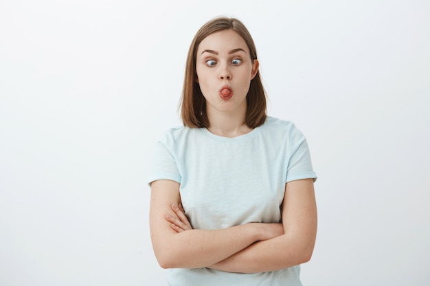 ¿puedes tocar la nariz con la lengua? retrato de mujer linda divertida e inmadura con pelo castaño corto entrecerrando los ojos haciendo muecas y jugando divirtiéndose sobre la pared gris