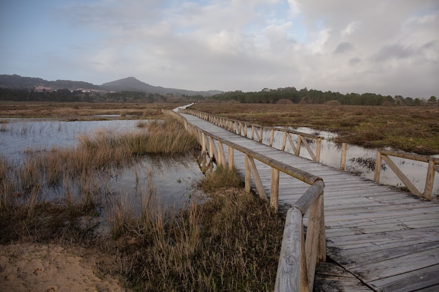 Puente de madera sobre un lago en un campo rodeado de colinas bajo un cielo nublado