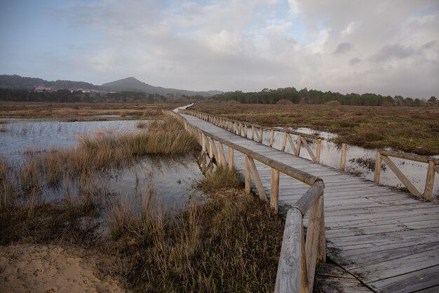 Puente de madera sobre un lago en un campo rodeado de colinas bajo un cielo nublado