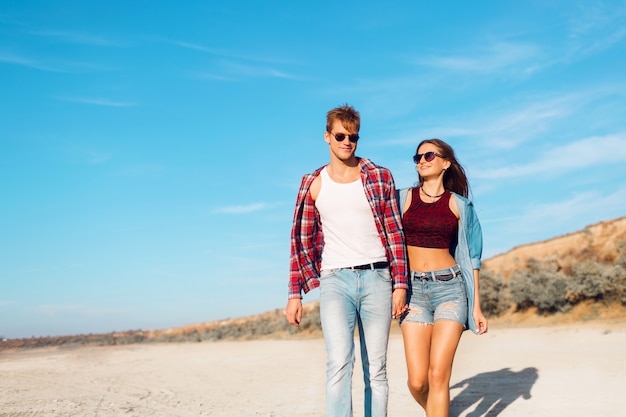 Puesta de sol, playa de arena, una amorosa pareja hipster camina abrazada en la playa desierta durante un día en la playa de vacaciones. Vistiendo ropa elegante de verano. Colores brillantes.