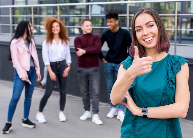 Foto gratuita pulgar que muestra femenino sonriente joven encima del gesto que mira la cámara mientras que sus amigos que se colocan empañan el fondo