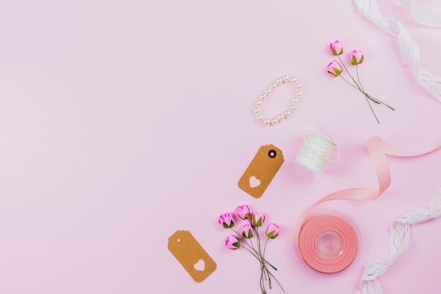 Foto gratuita pulsera de perlas; etiqueta; cinta; carrete de hilo; encaje y rosas artificiales sobre fondo rosa.