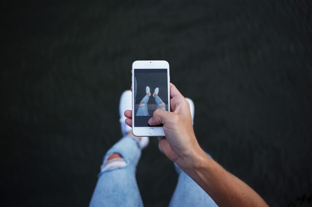 Punto de vista del hombre en jeans de mezclilla ajustados hipster de moda haciendo fotos de sus piernas y pies en zapatillas blancas en el teléfono inteligente para las redes sociales