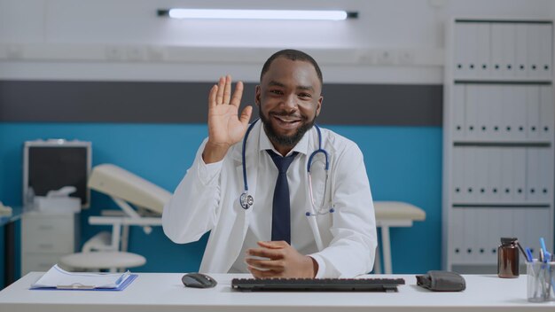 Punto de vista del médico especialista afroamericano sentado en el escritorio en la oficina del hospital explicando el tratamiento médico al paciente remoto durante la conferencia de la reunión de videollamada en línea. Concepto de telesalud