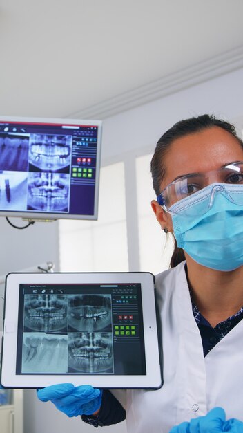 Punto de vista del paciente en el consultorio dental discutiendo el tratamiento de la cavidad de los dientes, dentista apuntando a la radiografía digital con tableta. Equipo de médicos que trabajan en la clínica estomatológica moderna, explicando el diente de radiografía