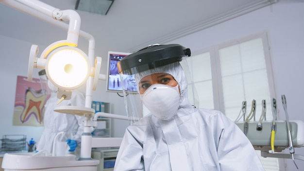 Punto de vista del paciente del dentista explicando el tratamiento de los dientes con traje protector covid en la nueva oficina estomatológica normal. Estomatólogo en equipo de seguridad contra coronavirus durante el control de atención térmica del paciente.