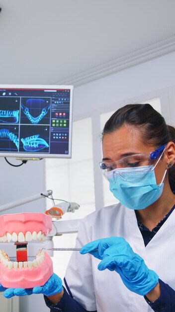 Punto de vista del paciente del dentista que muestra la forma correcta de limpiar los dientes en el consultorio dental con el accesorio de esqueleto médico de los dientes. Estomatólogo con máscara de protección durante el control del cuidado del calor