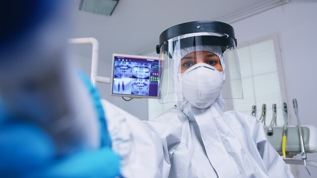 Punto de vista del paciente del dentista en traje de protección covid midiendo la temperatura en el consultorio dental moderno con nueva normalidad. Estomatólogo con equipo de seguridad contra el coronavirus durante el control de atención térmica de la persona.