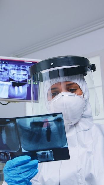 Punto de vista del paciente mirando al dentista en traje de ppe que muestra la imagen de rayos X en el consultorio dental. Especialista en estomatología vistiendo traje protector de materiales peligrosos contra el coroanvirus mostrando radiografía en la clínica con nueva normalidad