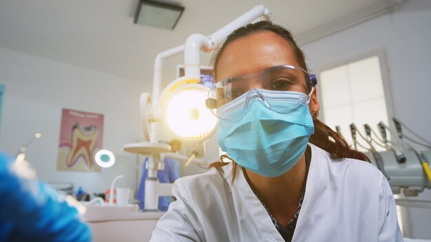 Punto de vista del paciente que visita la clínica dental para una cirugía que trata la masa afectada. Médico y enfermera trabajando juntos en la moderna oficina de ortodoncia, encendiendo la lámpara y examinando a la persona con máscara de protección.