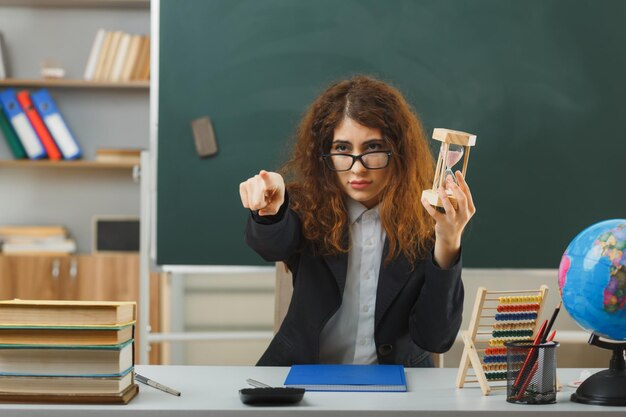 puntos de confianza en la cámara joven maestra con gafas sosteniendo un reloj de arena sentado en el escritorio con herramientas escolares en el aula