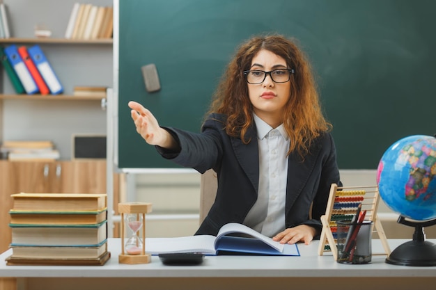 puntos estrictos al lado joven maestra con gafas sentada en el escritorio con herramientas escolares en el aula