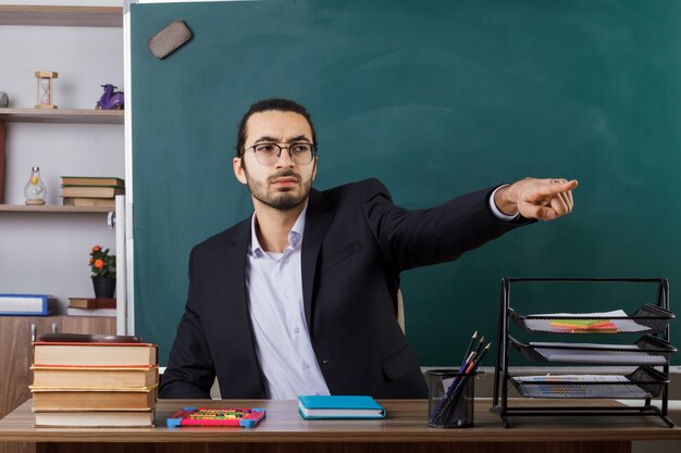 Puntos estrictos al lado del profesor con gafas sentado a la mesa con herramientas escolares en el aula