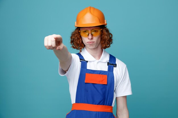 Puntos estrictos en cámara joven constructor hombre en uniforme aislado sobre fondo azul.