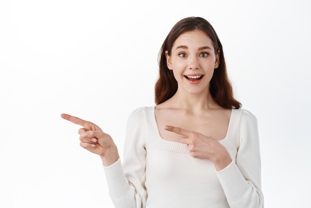 Los puntos femeninos jóvenes atractivos y positivos a un lado con una expresión alegre muestran algo increíble en un espacio en blanco aislado sobre un fondo blanco Concepto de publicidad