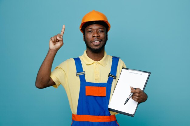 Puntos impresionados en el joven constructor afroamericano en uniforme sosteniendo portapapeles aislado sobre fondo azul.