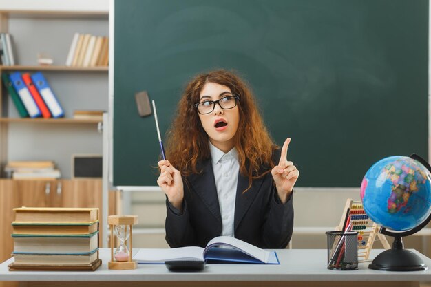 puntos impresionados en una joven maestra que usa anteojos sosteniendo un puntero sentado en el escritorio con herramientas escolares en el aula