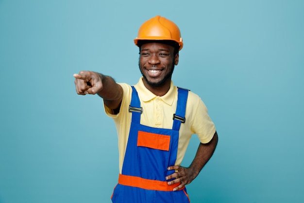 Puntos de risa al lado joven constructor afroamericano en uniforme aislado sobre fondo azul.