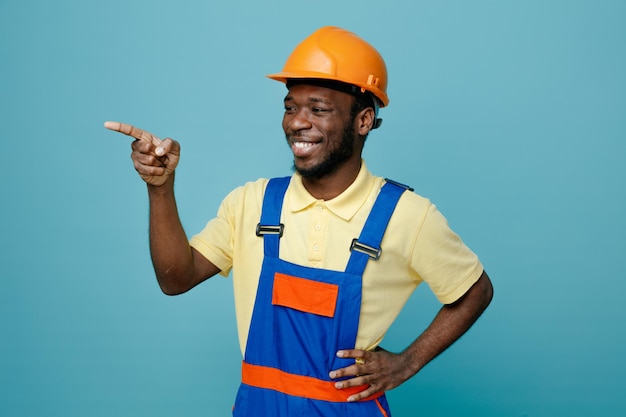 Puntos de risa al lado poniendo la mano en las caderas joven constructor afroamericano en uniforme aislado sobre fondo azul.