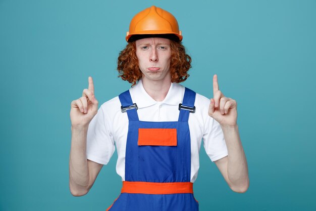 Puntos tristes en el hombre joven constructor en uniforme aislado sobre fondo azul.