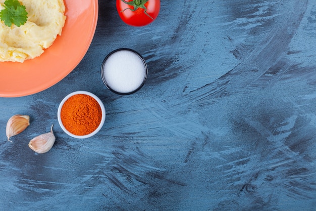 Puré de papas en un plato junto a verduras y tazones de especias, sobre la mesa azul.