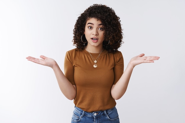 Y qué. Confundida preocupada joven mujer de cabello rizado oscuro encogiéndose de hombros con las manos extendidas hacia los lados levantar las cejas confundido expresar total incredulidad mirada desorientada perplejo discutiendo incapaz de entender lo que sucedió