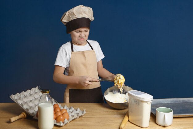 Que desastre. Niño decepcionado tratando de batir huevos con harina y leche mientras hace la masa por primera vez, pero tiene algún problema