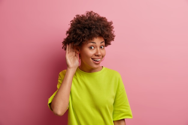 ¿Que dices? La mujer étnica positiva mantiene la mano cerca del oído para escuchar mejor los chismes, escucha la información privada, usa una camiseta informal, posa contra la pared rosa, escucha algo interesante