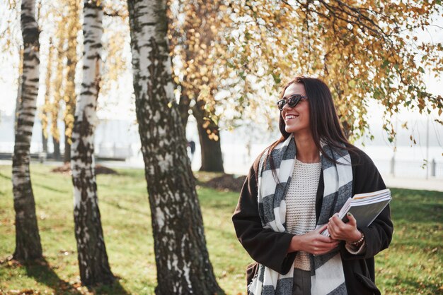 Que hermoso día. Joven morena sonriente en gafas de sol se encuentra en el parque cerca de los árboles y sostiene el bloc de notas