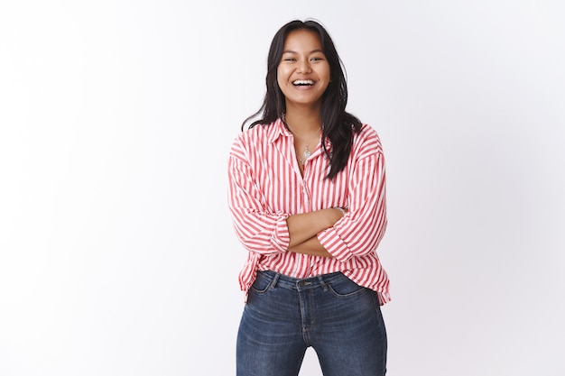 Qué hermoso día. Retrato de alegre, optimista y energizada linda chica malaya en blusa a rayas rosa divirtiéndose bromeando, teniendo una conversación divertida, sonriendo y riendo a la cámara sobre la pared blanca