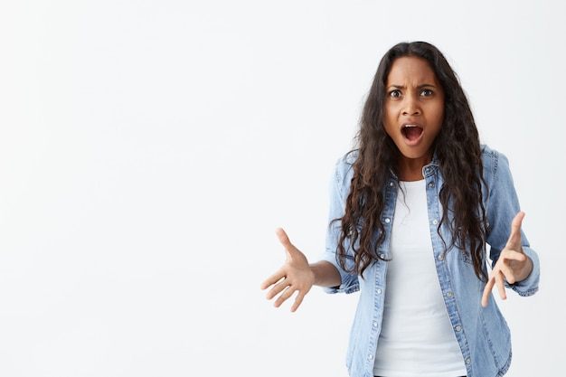 ¡¿Qué?! Insatisfecha mujer de piel oscura con cabello ondulado negro encogiéndose de hombros en gesto de interrogación, gesticulando activamente y manteniendo la boca abierta, con una mirada confundida y enojada.